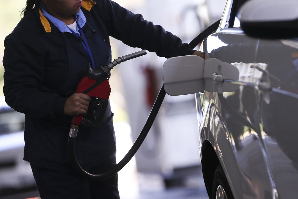 Mulher de uniforme azul segura bomba de combustíveis vermelha e abre a tampa do tanque de automóvel branco. Fiscais do Procon-DF fazem vistoria em postos de combustíveis de Brasília para checar as alterações dos preços finais, após aumento dos impostos federais PIS-Cofins (Foto: Marcelo Camargo/Agência Brasil)