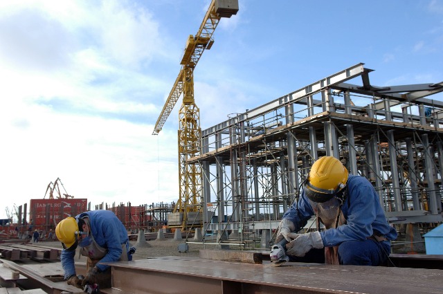 Na imagem: Dois trabalhadores serram vigas de ferro (com macacão azul, capacete amarelo, luvas e óculos de proteção), em canteiro de obra no Estaleiro Rio Grande, da Ecovix; ao fundo, grande estrutura metálica em construção (Foto: Stéferson Faria/Agência Petrobras)