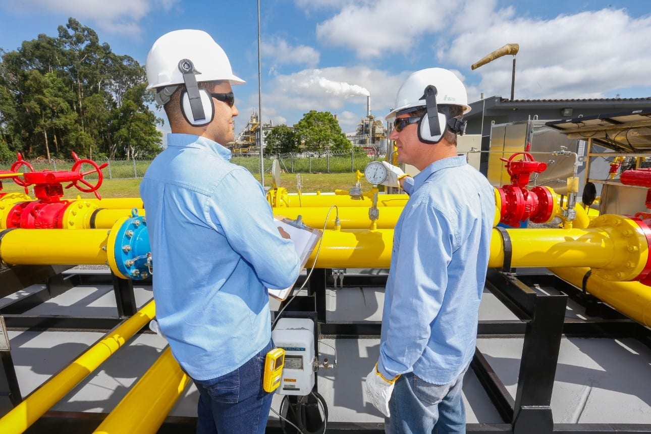 Citygate (ponto de entrega de gás) da Compagas, distribuidora de gás natural do Paraná; Rede de gasodutos nas cores amarela e vermelha, com dois trabalhadores realizando inspeção e conversando; vestem uniforme azul, capacete branco e óculos escuros, equipamentos de proteção individual (Foto: Divulgação)