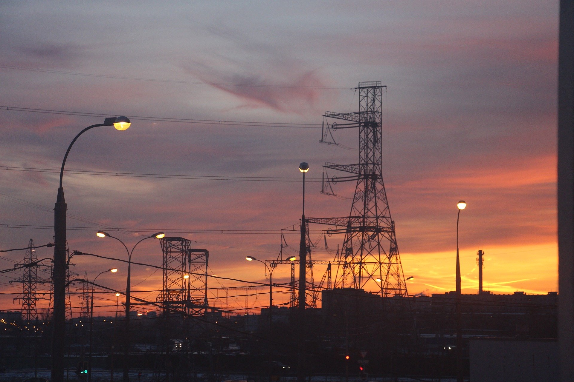 Pôr do sol ao fundo de torres com linhas de transmissão de energia, com alguns postes com luzes acesas (Foto Inna/Pixabay)