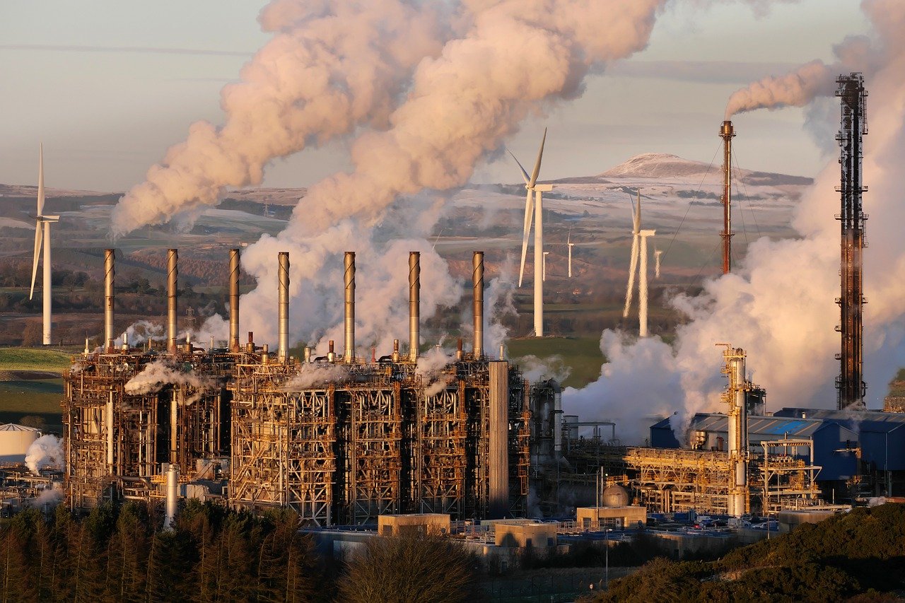 Setor de energia em desalinho com limite de 2°C para aquecimento global. Na imagem: Chaminés de indústria de petróleo emitindo grande volume de gases de efeito estufa, gerando uma densa fumaça branca e, ao fundo, turbinas eólicas (Foto: Emphyrio/Pixabay)