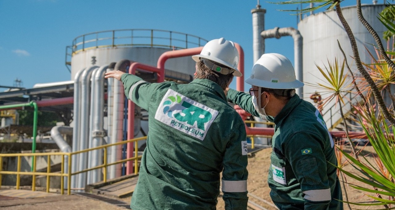 3R fecha, com Bahiagás, seu primeiro contrato para venda de gás natural. Na imagem: Dois trabalhadores em planta da 3R Petroleum. Vestem macacão verde escuro com sinalizações de segurança e logo escrito "3R Petroleum", com destaque, nas costas. Usam capacete branco. Um deles aponta para as instalações com o braço esquerdo estendido (Foto: Divulgação)