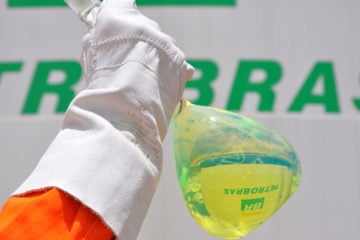 Petrobras: Biorrefino vai exigir infraestrutura com dutos dedicados para biocombustíveis. Na imagem: Mão, com luva de proteção branca, segura frasco de laboratório, de vidro, contendo amostra de diesel com teor renovável, da Petrobras, um líquido esverdeado e translúcido; ao fundo, tanque de armazenamento de combustíveis (Foto: Michel Chedid/Petrobras)