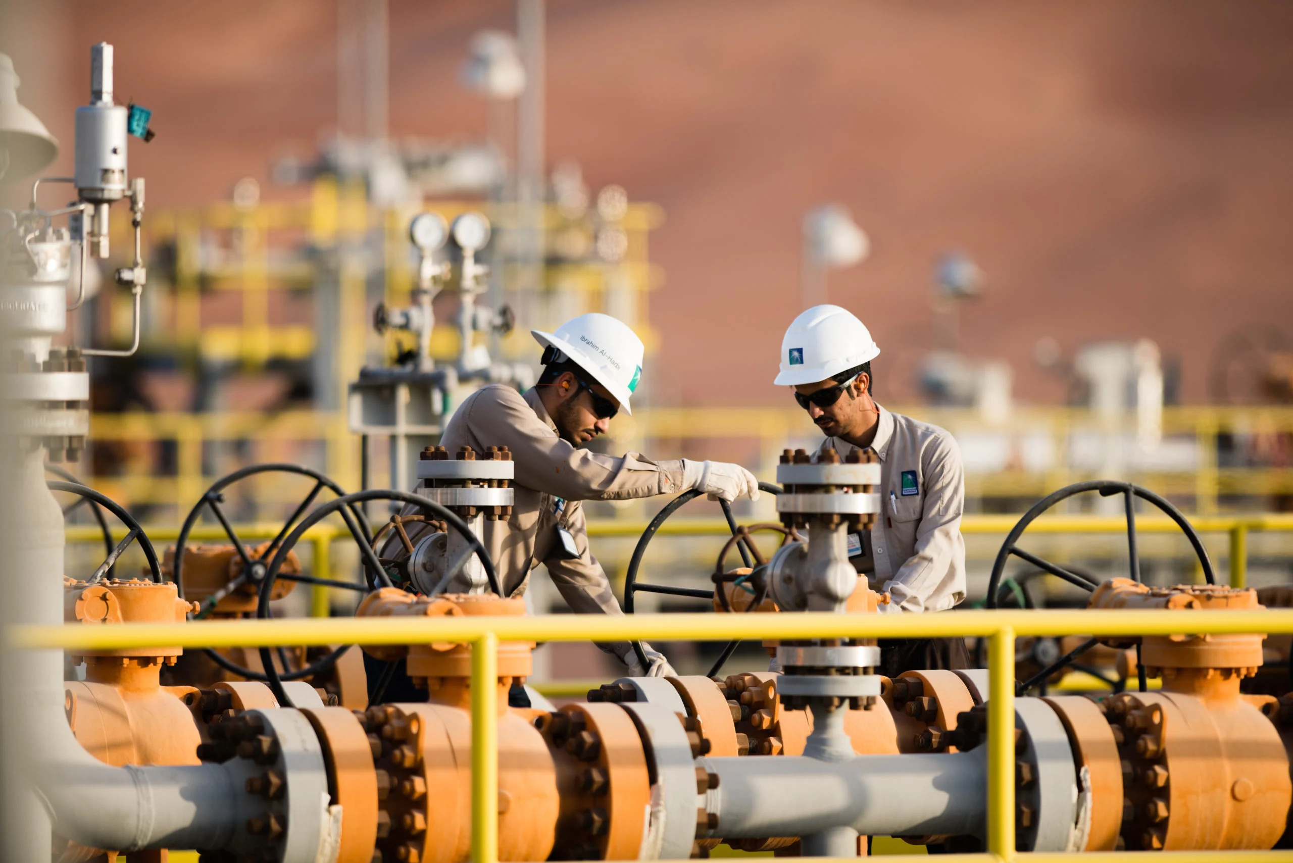 Dois trabalhadores, com uniformes cinza e capacete de proteção branco, operam instalações em gasoduto (Foto Divulgação Saudi Aramco)