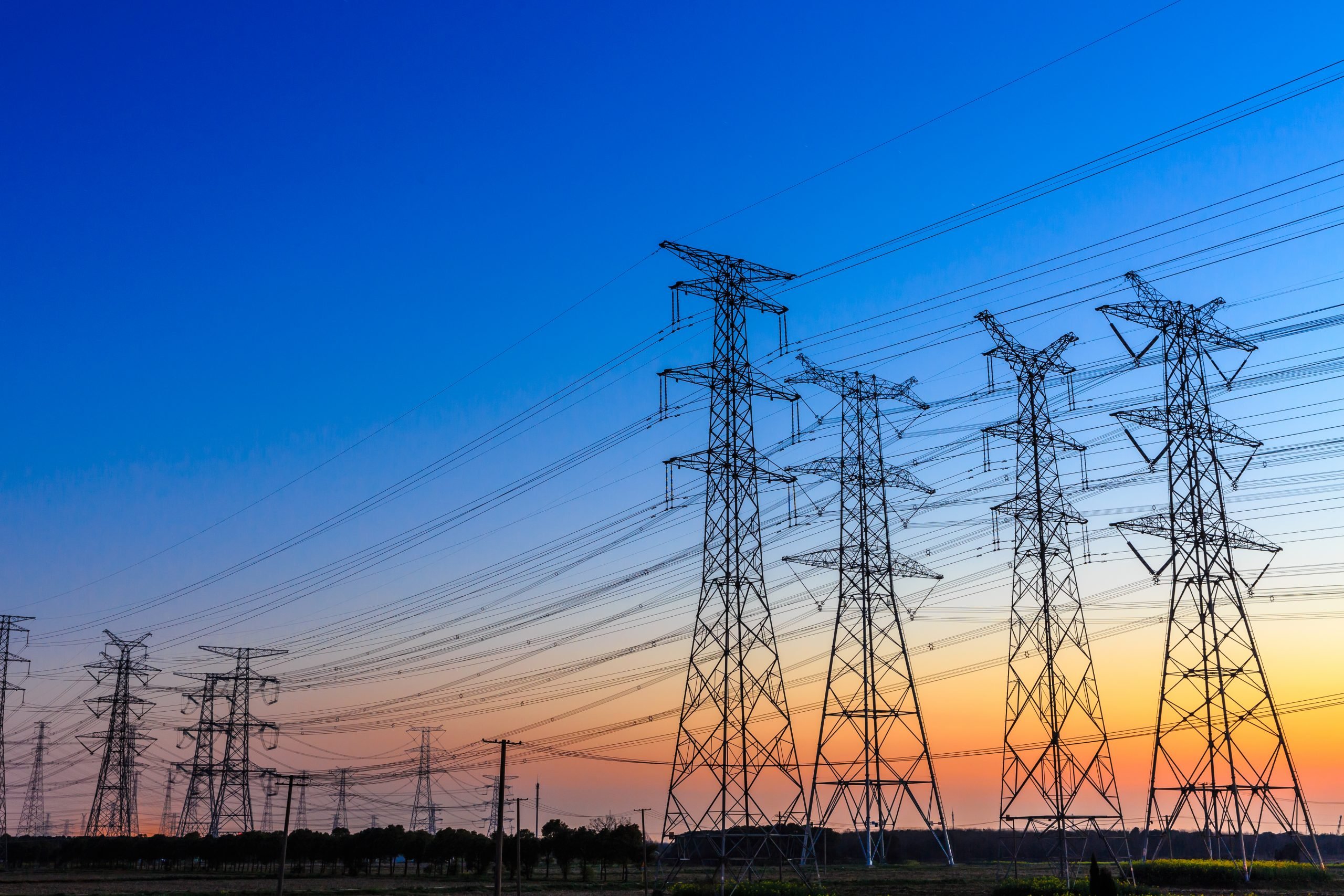 Mercado livre de energia bate recorde de novos consumidores no 1º semestre de 2023. Na imagem: Foto à contraluz de grandes torres com linhas de transmissão de energia ,sob céu azul com pôr do sol ao fundo (Foto: iStock)