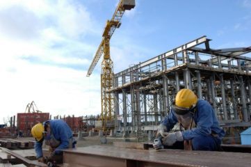 Plataformas de petróleo, gás natural e refino vão aquecer estaleiros e indústria no Brasil. Na imagem: Dois trabalhadores serram vigas de ferro (com macacão azul, capacete amarelo, luvas e óculos de proteção), em canteiro de obra no Estaleiro Rio Grande, da Ecovix; ao fundo, grande estrutura metálica em construção (Foto: Stéferson Faria/Agência Petrobras)