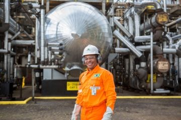 A Petrobras convocará 2.170 aprovados de concursos e planeja um novo concurso técnico com 458 vagas até o final de 2023. Na imagem: Trabalhador sorrindo, com uniforme laranja e equipamentos de proteção, em frente a rede de dutos e instalações metálicas, na cor prata, em operação de Petrobras (Foto: Agência Petrobras)