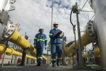 Petrobras fecha contrato de R$ 56 bilhões com a Comgás para 2024. Na imagem: Trabalhadores em planta de gasodutos da Comgás; Um homem e uma mulher caminham com uniformes azuis com faixas de proteção amarelas fluorescente, capacetes e óculos de proteção e protetores auriculares (Foto: Divulgação)