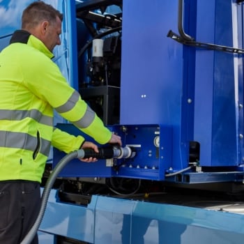 Estratégia brasileira para hidrogênio do governo federal prevê hubs até 2035; transporte pesado pode alavancar demanda. Na imagem: Caminhão elétrico azul da Volvo conectado a carregador elétrico por trabalhador com uniforme amarelo. Volvo apresenta na Europa e EUA caminhão com células de combustível a hidrogênio, com zero emissões (Foto: Divulgação Volvo)