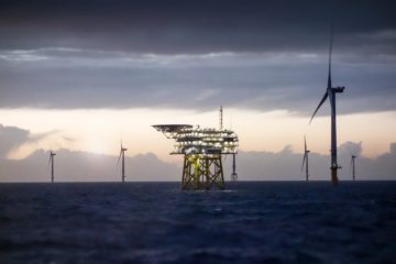 A crise climática é uma realidade e o Brasil precisa aproveitar seus enormes potenciais na energia. Na imagem: Plataforma offshore de petróleo e gás com turbinas eólicas associadas, durante pôr do sol, com mar azul bem escuro e céu encoberto com nuvens acinzentadas (Foto: Charlie Chesvick/Getty Images)