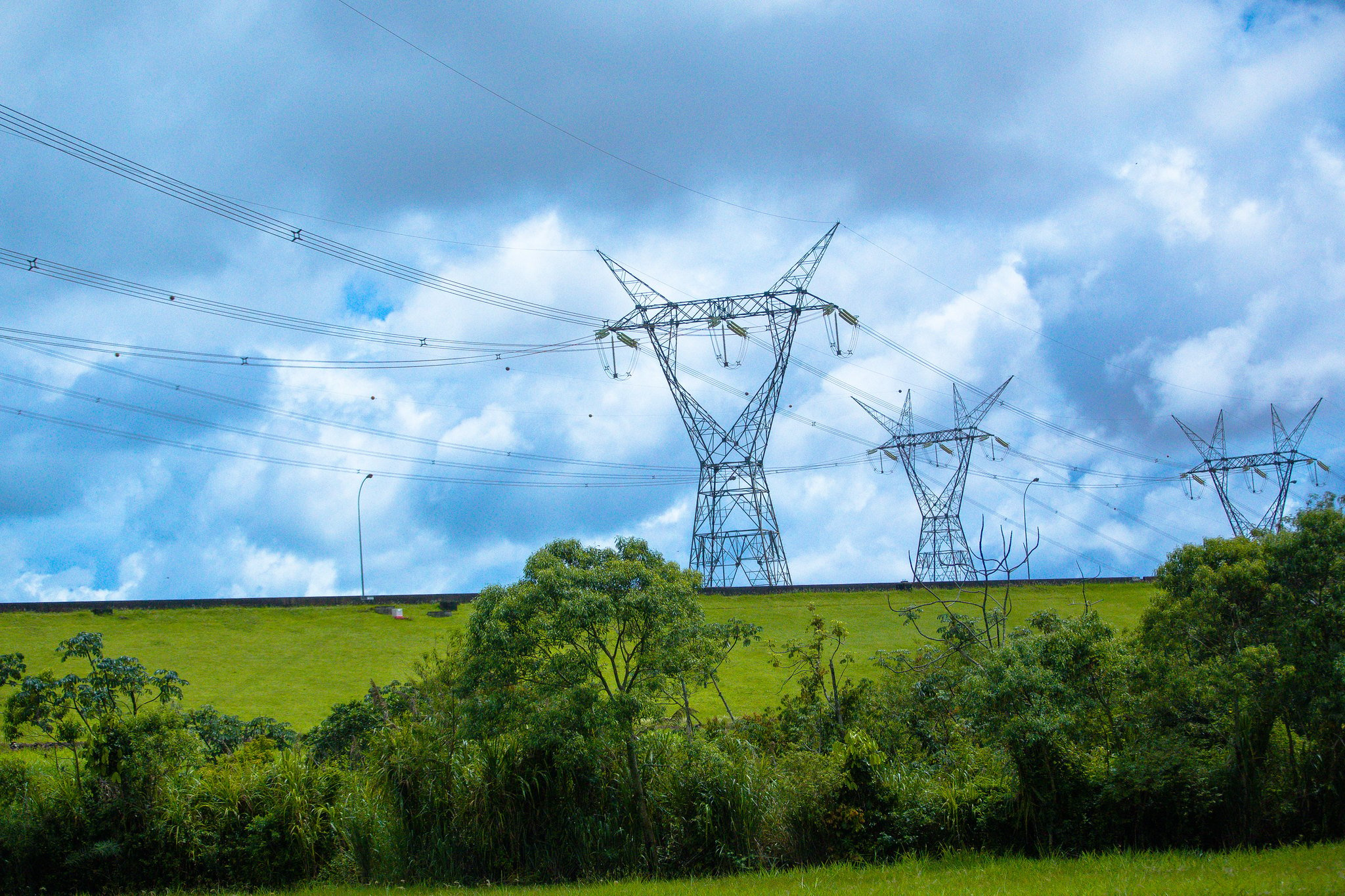 Falha em linhas de transmissão interrompe fornecimento de energia elétrica no Rio de Janeiro. Na imagem: Linha de transmissão com cabos de alta tensão da usina de Itaipu, em Foz do Iguaçu (PR), em 16/1/2018; Foto de torres ao fundo sobre pastagem verde com alguns arbustos, em primeiro plano, e céu encoberto de nuvens brancas acinzentadas (Foto: Divulgação)