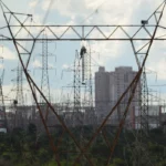 Aneel anuncia que conta de luz terá bandeira amarela para o mês de julho. Na imagem: Linhas de transmissão de energia em área urbana, com prédios ao fundo (Foto: Agência Brasil)