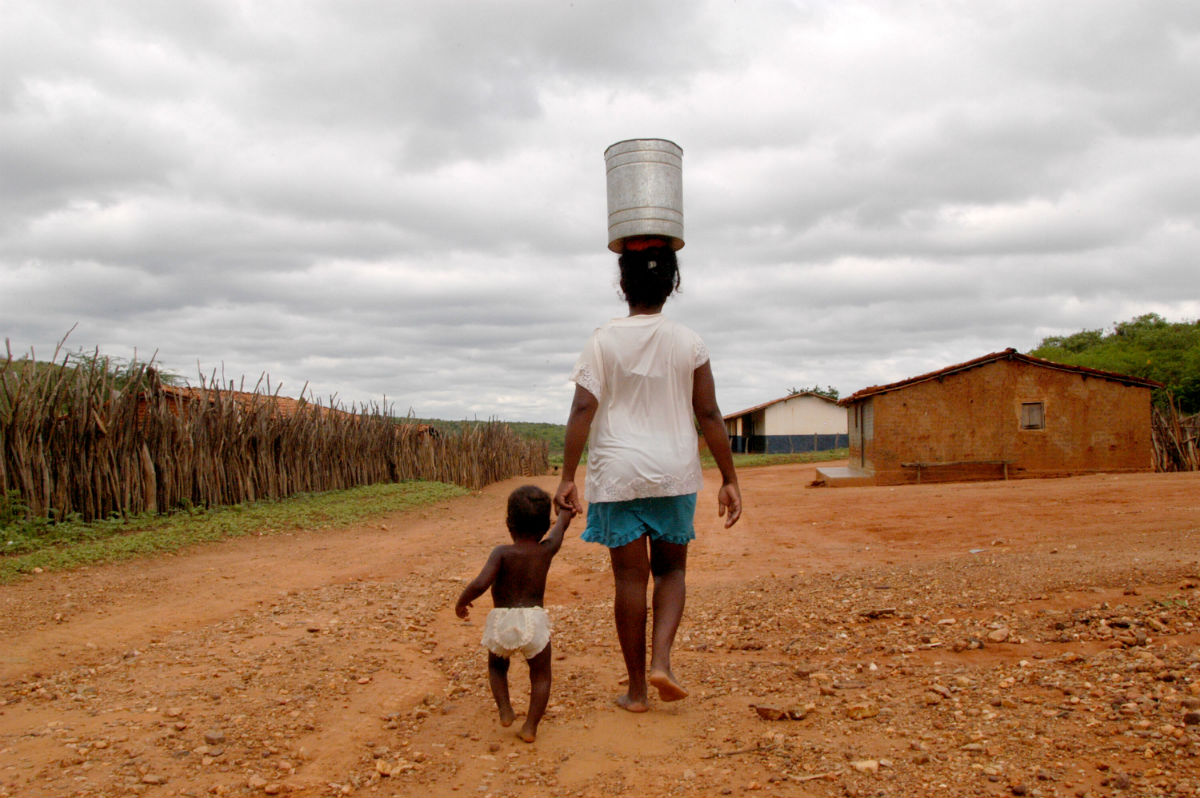 Vamos falar sobre as mulheres em situação de pobreza energética sem acesso a energia elétrica e combustível limpo para cozinhar. Na imagem: Mulher carrega lata d'água na cabeça no Nordeste brasileiro e segura uma criança pela mão caminhando sobre chão de terra (Foto: ClimaInfo)