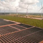 Na imagem: Vista de usina híbrida eólica e solar fotovoltaica, com inúmeras fileiras de módulos fotovoltaicos, em primeiro plano, e aerogeradores, ao fundo, à direita (Foto Divulgação Voltalia)