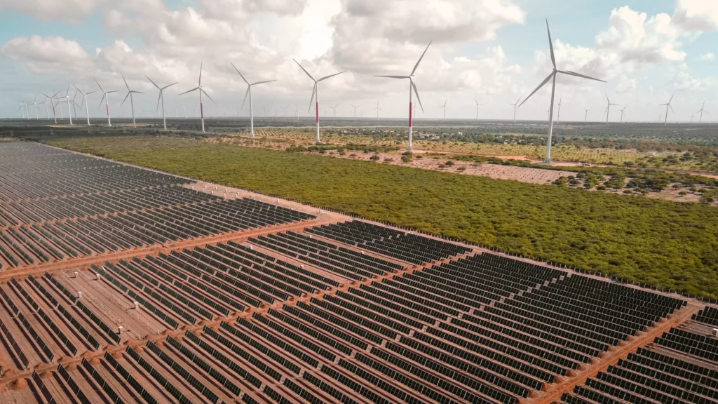 Vista de usina híbrida eólica e solar fotovoltaica, com inúmeras fileiras de módulos fotovoltaicos, em primeiro plano, e aerogeradores, ao fundo, à direita (Foto Divulgação Voltalia)