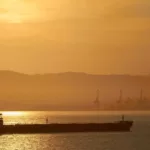 Navio-tanque transporta petróleo com céu em tons de amarelo com pôr do sol, e montanha, ao fundo (Foto Creative Commons)