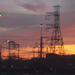 Pôr do sol ao fundo de torres com linhas de transmissão de energia, com alguns postes com luzes acesas (Foto Inna/Pixabay)
