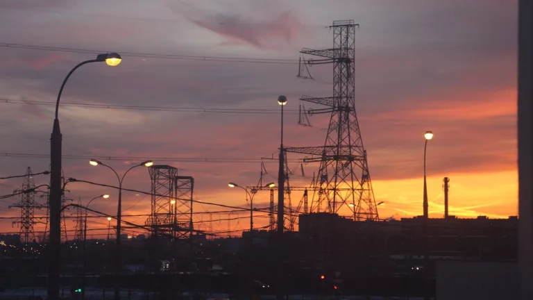 Pôr do sol ao fundo de torres com linhas de transmissão de energia, com alguns postes com luzes acesas (Foto Inna/Pixabay)