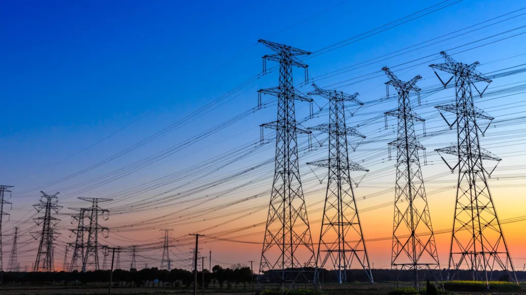 Foto à contraluz de grandes torres com linhas de transmissão de energia, sob céu azul com pôr do sol ao fundo (Foto iStock)
