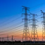 Foto à contraluz de grandes torres com linhas de transmissão de energia, sob céu azul com pôr do sol ao fundo (Foto iStock)