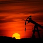 Foto à contraluz de cavalo-de-pau para a exploração de petróleo em terra, com sol se pondo ao fundo e céu alaranjado (Foto Zbynek Burival/Unsplash)