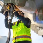 Técnico de manutenção de aviação, com colete de segurança amarelo fosforescente, carrega combustível de aviação sustentável no Boeing ecoDemonstrator 2021, com mangueira conectada verticalmente na parte de baixo da aeronave (Foto Divulgação Boeing)