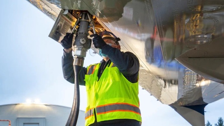 Técnico de manutenção de aviação, com colete de segurança amarelo fosforescente, carrega combustível de aviação sustentável no Boeing ecoDemonstrator 2021, com mangueira conectada verticalmente na parte de baixo da aeronave (Foto Divulgação Boeing)