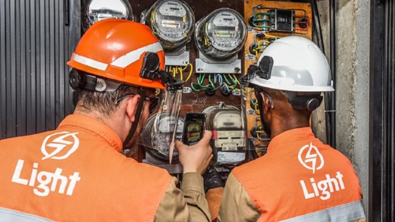 Técnicos da Light, um homem branco e um negro uniformizados, realizam medições e fotografam relógio de energia (Foto Divulgação)