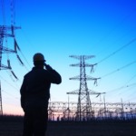 Foto &agrave; contraluz de operador do setor el&eacute;trico ao telefone e, ao fundo, linhas de transmiss&atilde;o, com c&eacute;u azul (Foto Divulga&ccedil;&atilde;o)