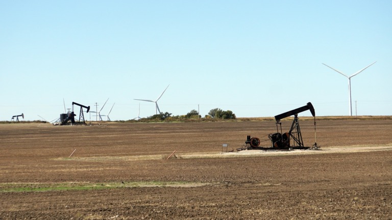 Cavalos-de-pau (bomba 'pumpjack') para a produção onshore de petróleo, em Oklahoma (EUA), com turbinas eólicas ao fundo (Foto Mike/RJA1988/Pixabay)