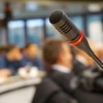 Microfone direcional em p&uacute;lpito vazio, em primeiro plano, e, ao fundo, imagem desfocada de plateia em bancadas de sala de confer&ecirc;ncias(Foto Foto Florian Pircher/Pixabay)